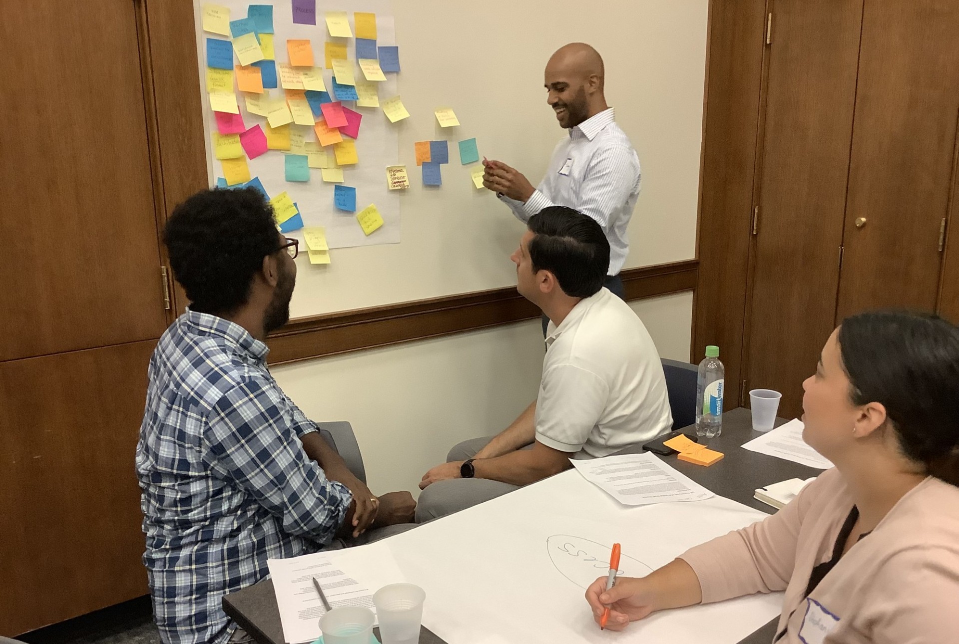 Two men and a woman sitting and looking at a whiteboard covered with sticky notes on the left half, with a man standing to the right of the board